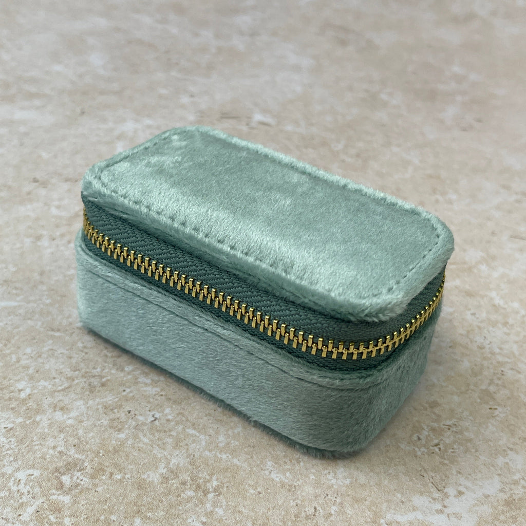 A small rectangular mint green velvet zipper case with a gold zipper, resting on a textured beige stone surface.